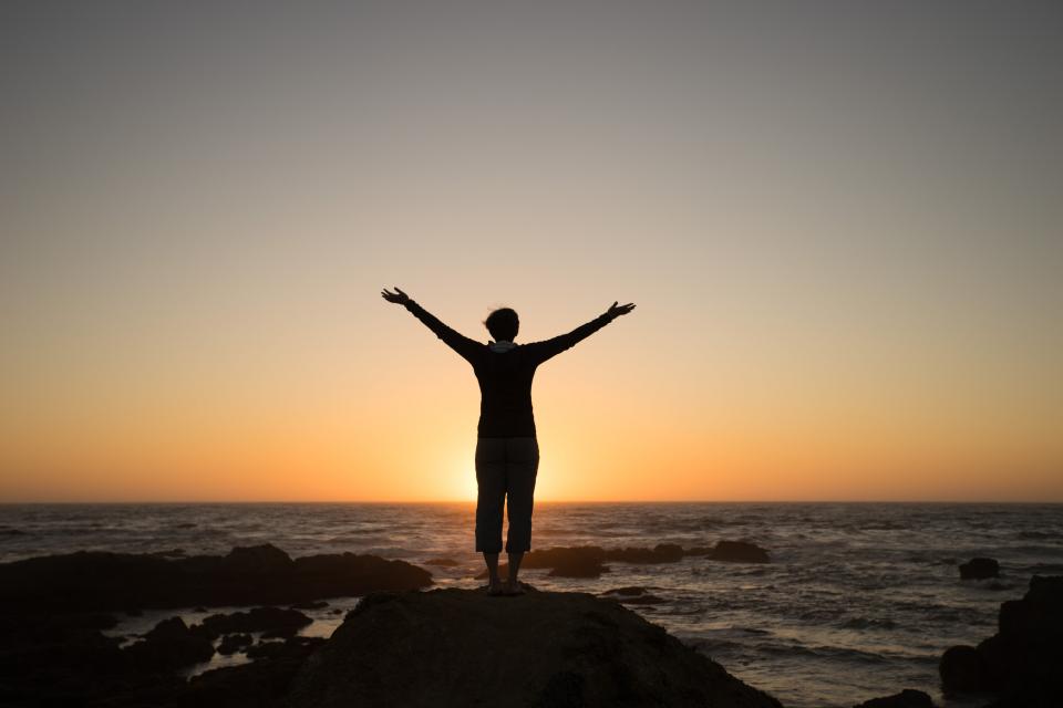 Woman stands arms stretched from a rock overlooking a sunset over water