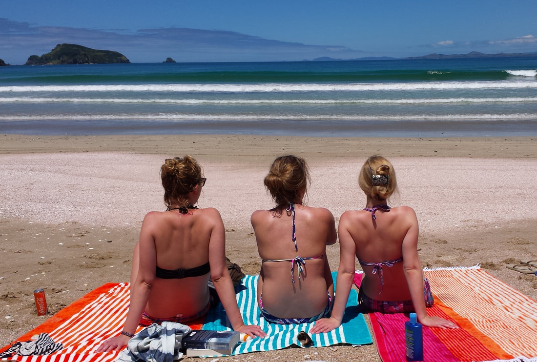 Three women facing away from the camera sitting on colourful beach towels overlooking the beach and the ocean in the Bay of Islands, New Zealand 