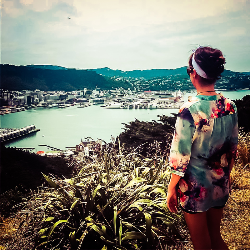 Picture of a woman standing on the top of a hill and looking down at a city and the sea in New Zealand