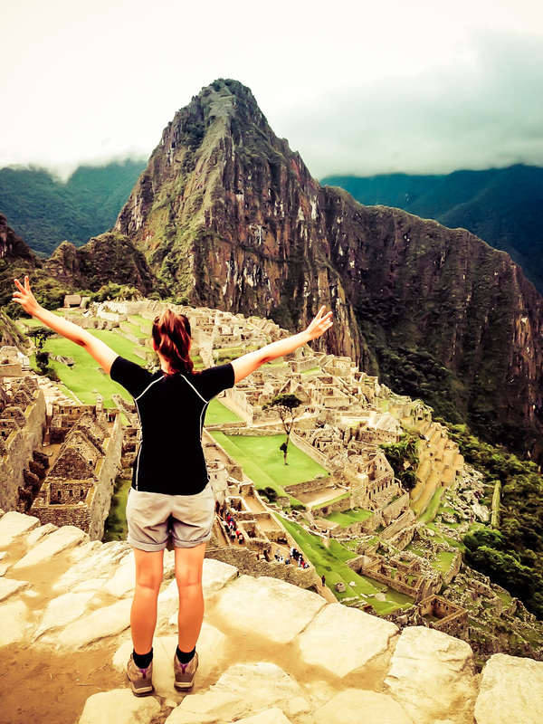 Picture  of a woman standing on top of a hill overlooking Machu Picchu in South America with her hand up in the air 