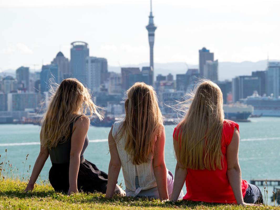 Picture of three woman sitting on a grassy hill overlooking the sea and skyline of Auckland in New Zealand 