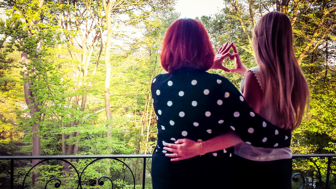 Picture of two women overlooking trees from a balcony forming a heart shape with their hands