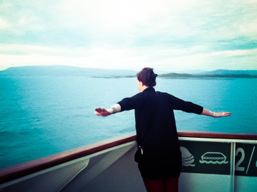 Picture of a woman standing at the back of a boat with outstretched arms looking out to sea
