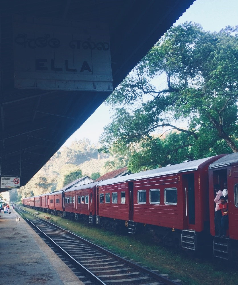 A red train departing the train station in Ella, Sri Lanka with passengers standing in the doorways of the train and on the platform 