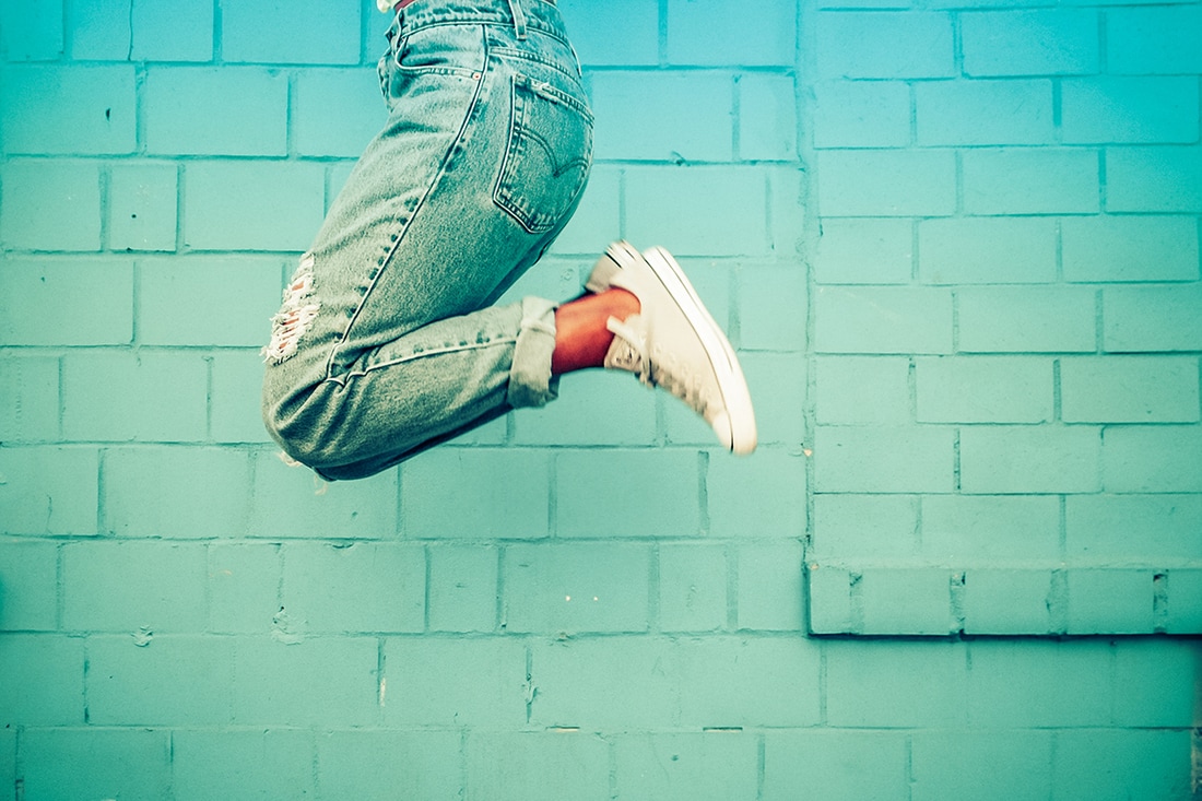 Picture of a woman jumping up in the air in front of a turquoise coloured wall 