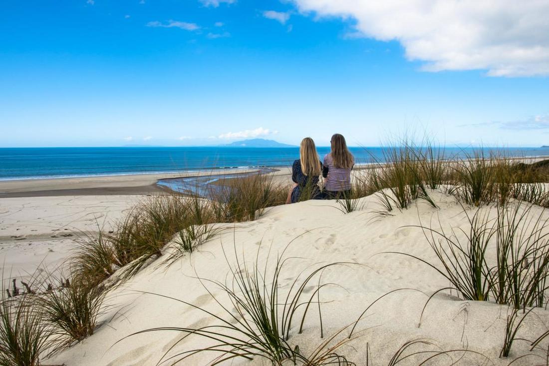 Picture of two woman sitting on a sand dune looking at the beach and the ocean in Mangawhai, New Zealand 