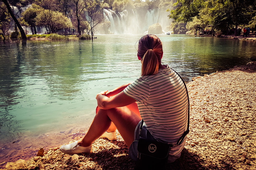 Picture of a woman sitting on the shore of a lake looking at a waterfall 