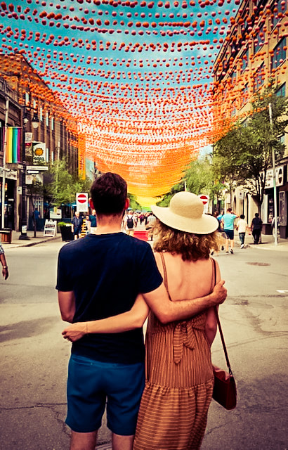 A couple embracing each other facing away from the camera as they look ahead of them at a street that is framed by colourful lanterns in Montreal