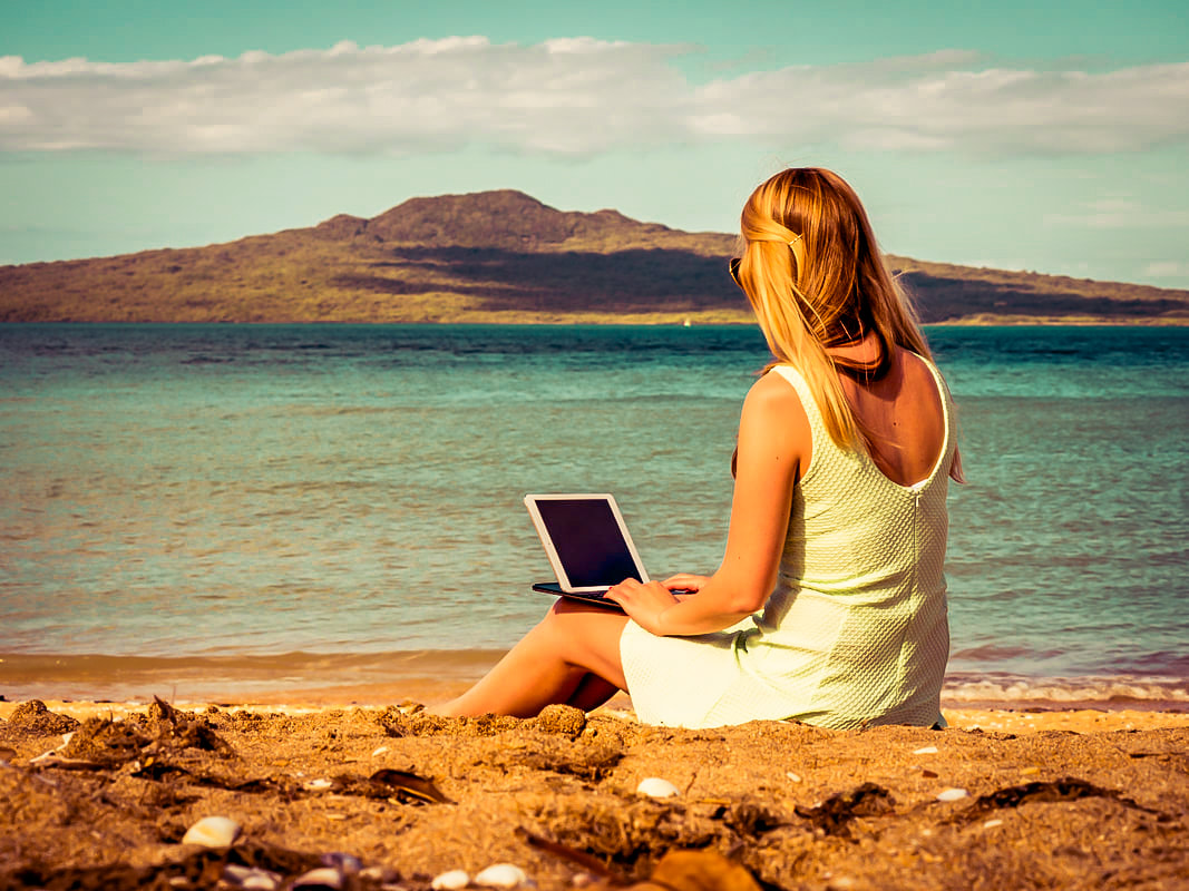 Picture of a woman sitting on a beach, working remotely on her laptop, facing away from the camera, overlooking the ocean and a volcano in Auckland, New Zealand 