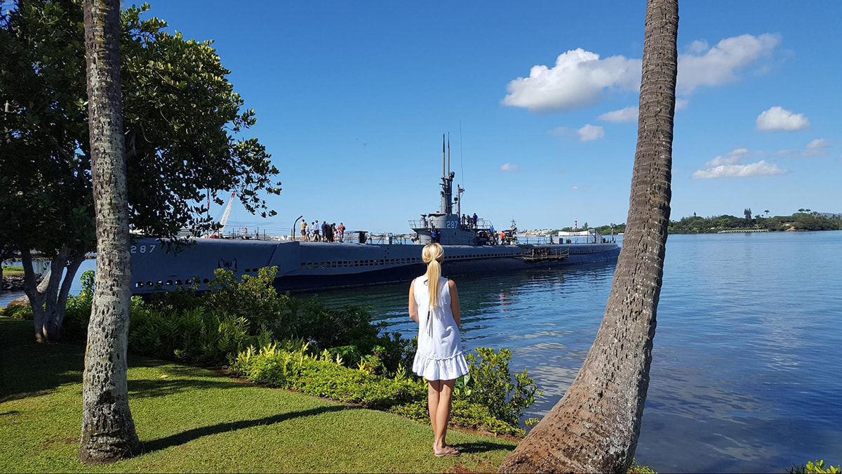 Woman standing underneath palm trees looking out to a ship at the pearl harbour grounds in Hawaii